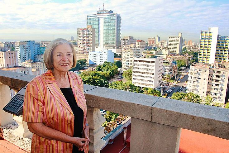 Ursula Männle steht auf einem Balkon, hoch über den Dächern Havannas. Im Hintergrund höhere Wohngebäude und ein gläsern verschalter Büroturm.