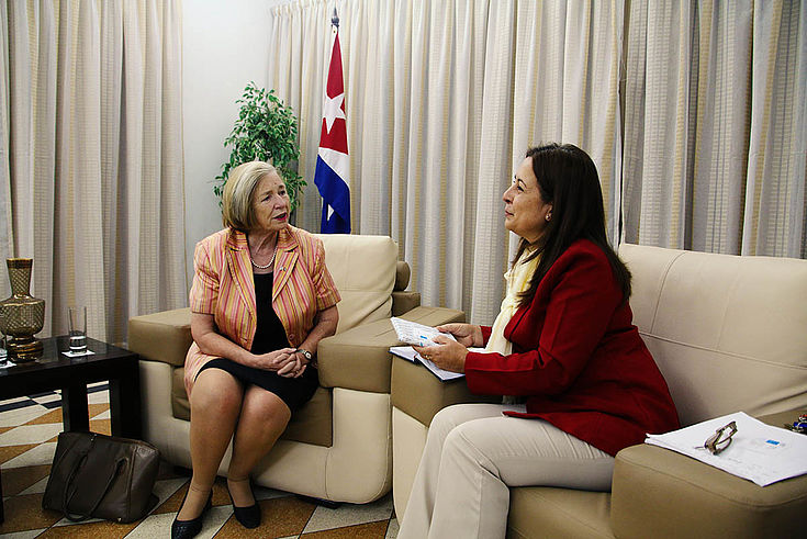 Ursula Männle im Gespräch mit Inalvis Bonachea auf zwei gemütlichen Sesseln. Im Hintergrund steht eine Flagge Kubas.