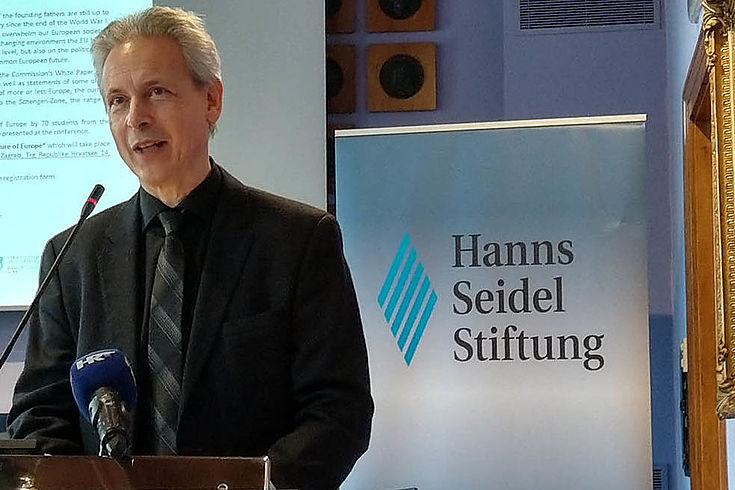 Ein Mann steht an einem Podium und spricht gerade. Dahinter eine Leinwand und ein Roll-Up der Hanns-Seidel-Stiftung.