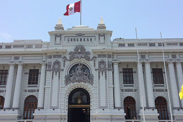 Weißes staatstragendes Gebäude mit peruanischer Flagge vor blauem Himmel.