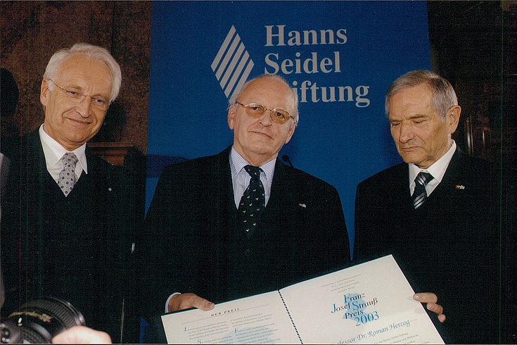 drei ältere Männer in dunklem Anzug vor Logo der Hanns-Seidel-Stiftung