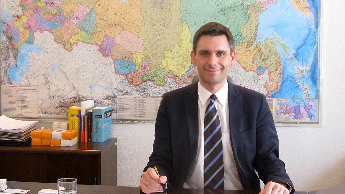 Herr Dresel sitzt freundlich lächelnd hinter seinem Schreibtisch, einen Füller in der Hand über einem Blatt Papier. Hinter ihm eine Karte Russlands an der Wand seines Büros.