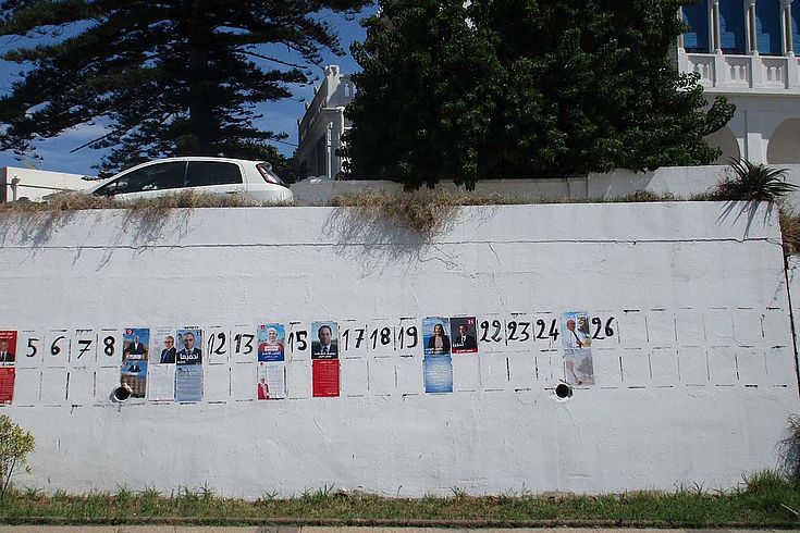 Eine weiße Mauer mit vorgezeichneten Flächen für Wahlplakate. Im Hintergrund Bäume.