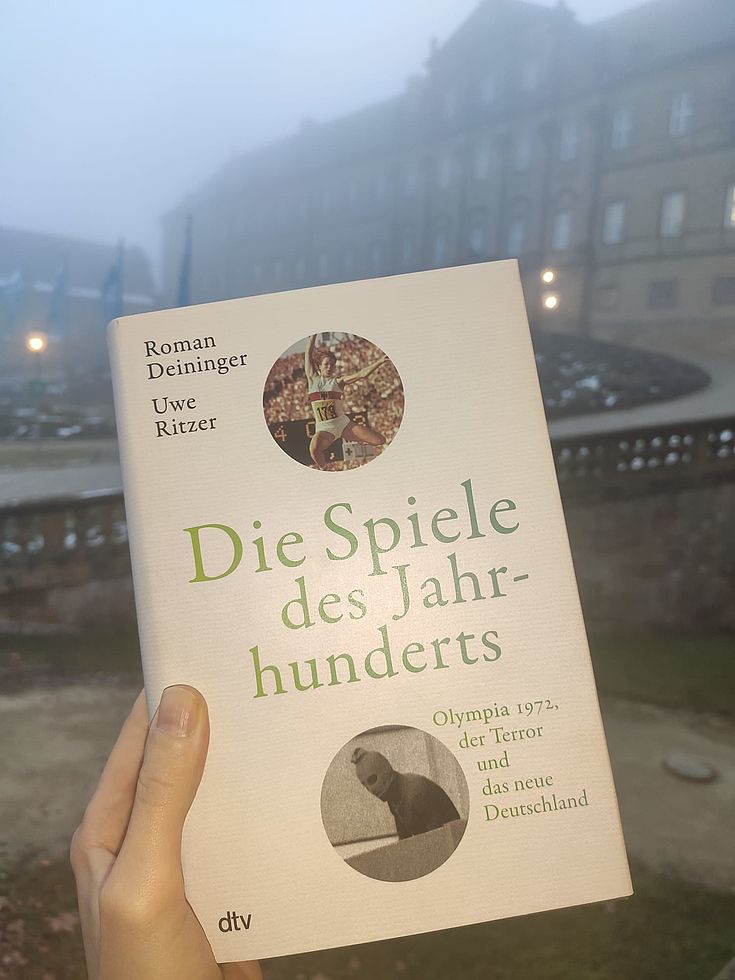 Das Buch wird von einer Hand ins Bild gehalten. Im Hintergrund der Innenhof des Klosters Banz. 