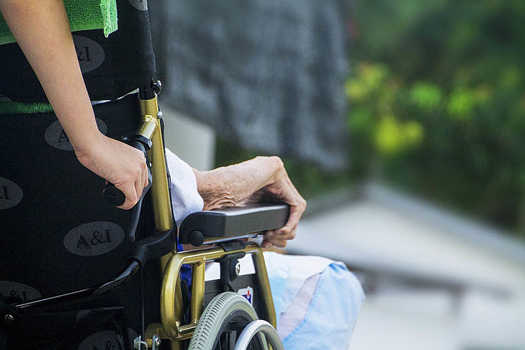 Ein Rollstuhl von hinten, mit einer Hand eines alten Menschen auf der Lehne und einer Hand eines jungen Menschen, der den Rollstuhl schiebt.