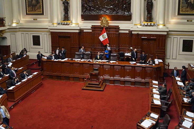 Blick in den peruanischen Kongresssaal mit hufeisenförmig angeordneten Sitzen und rotem Teppich. Vorne die Landesflagge vor einem Podium.